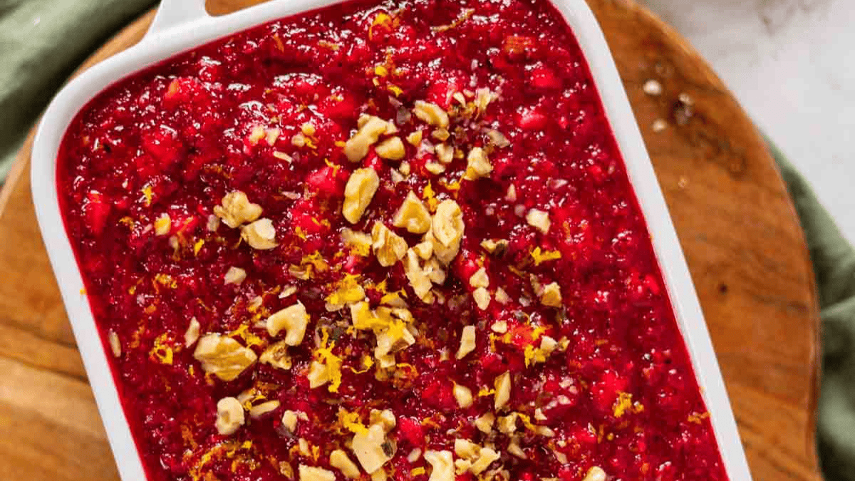 A white baking dish filled with bright red cranberry salad topped with chopped walnuts and orange zest, placed on a wooden surface.
