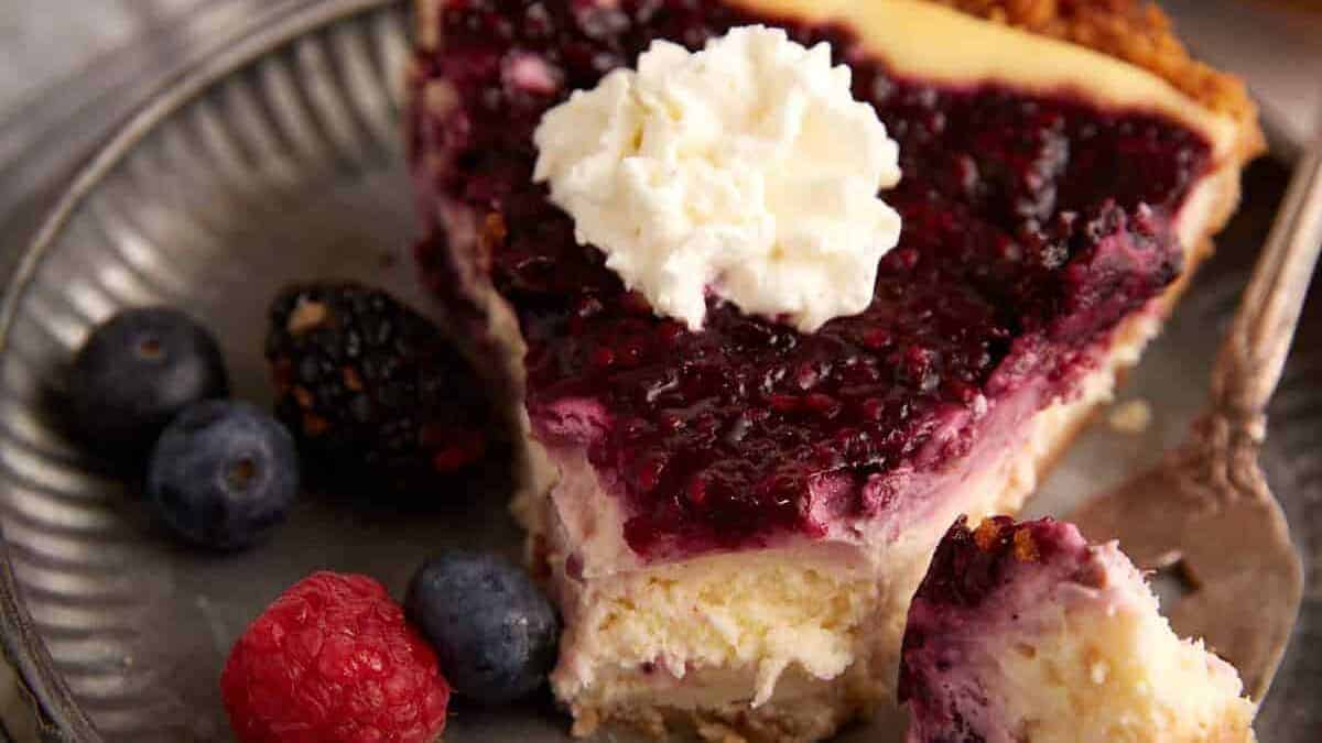 A slice of berry cheesecake topped with whipped cream sits on a silver plate, accompanied by fresh blackberries, blueberries, and a raspberry, with a fork holding a bite of the dessert.