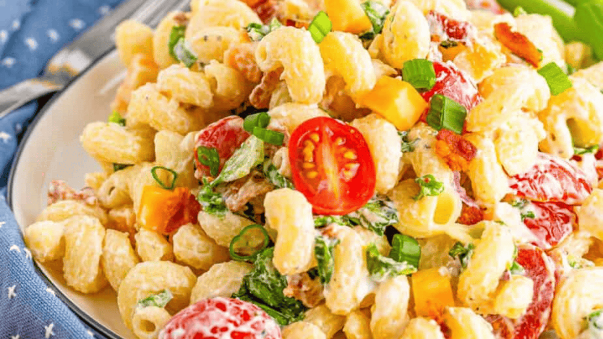 A close-up of a creamy pasta salad with cavatappi noodles, cherry tomatoes, green onions, cheddar cheese cubes, lettuce, and crispy bacon bits on a plate.