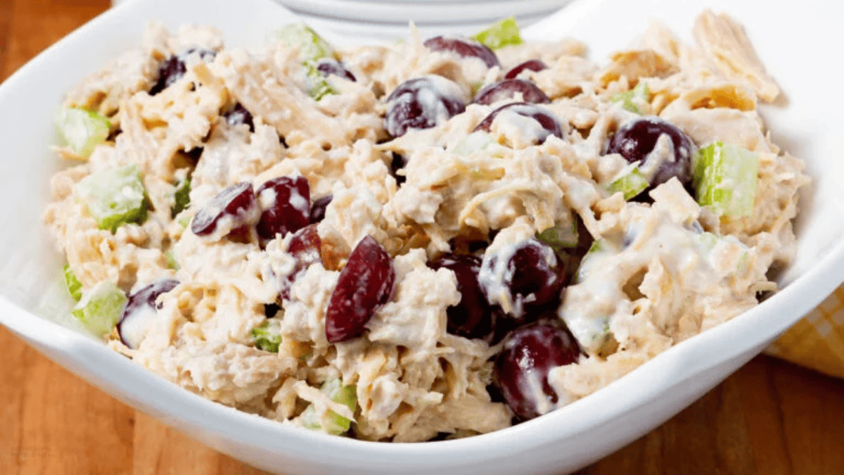 A bowl of chicken salad with pieces of chicken, red grapes, chopped celery, and a creamy dressing, served in a white dish on a wooden table.