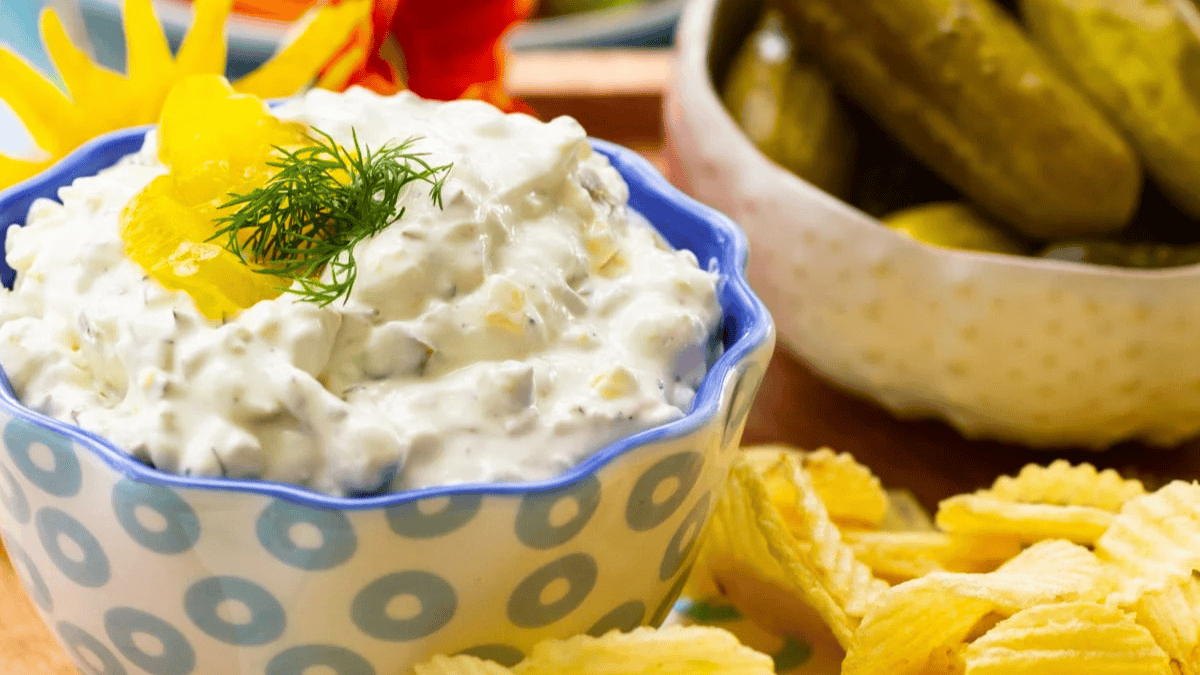 A bowl of creamy dip garnished with dill and yellow peppers, surrounded by ridged potato chips. In the background, there is a bowl of whole pickles and a decorative flower.