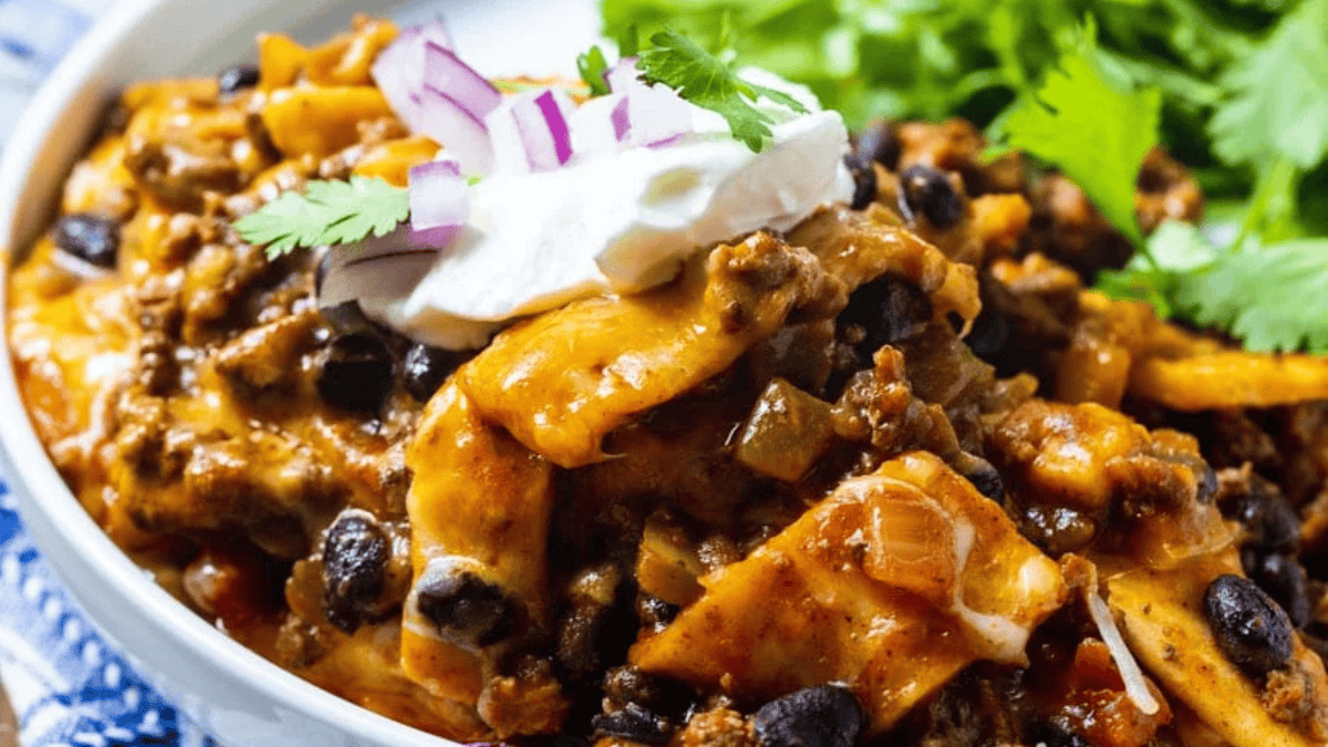 A close-up of a cheesy Tex-Mex casserole with ground beef, black beans, tortilla strips, melted cheese, and spices, topped with sour cream, chopped red onions, and cilantro, served with a side of fresh greens.
