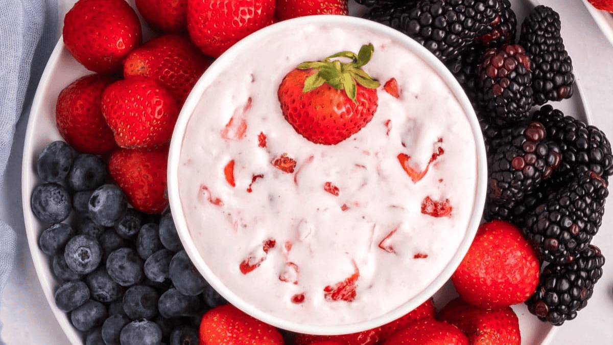 A bowl of creamy fruit dip topped with a strawberry, surrounded by fresh strawberries, blackberries, and blueberries on a white plate.