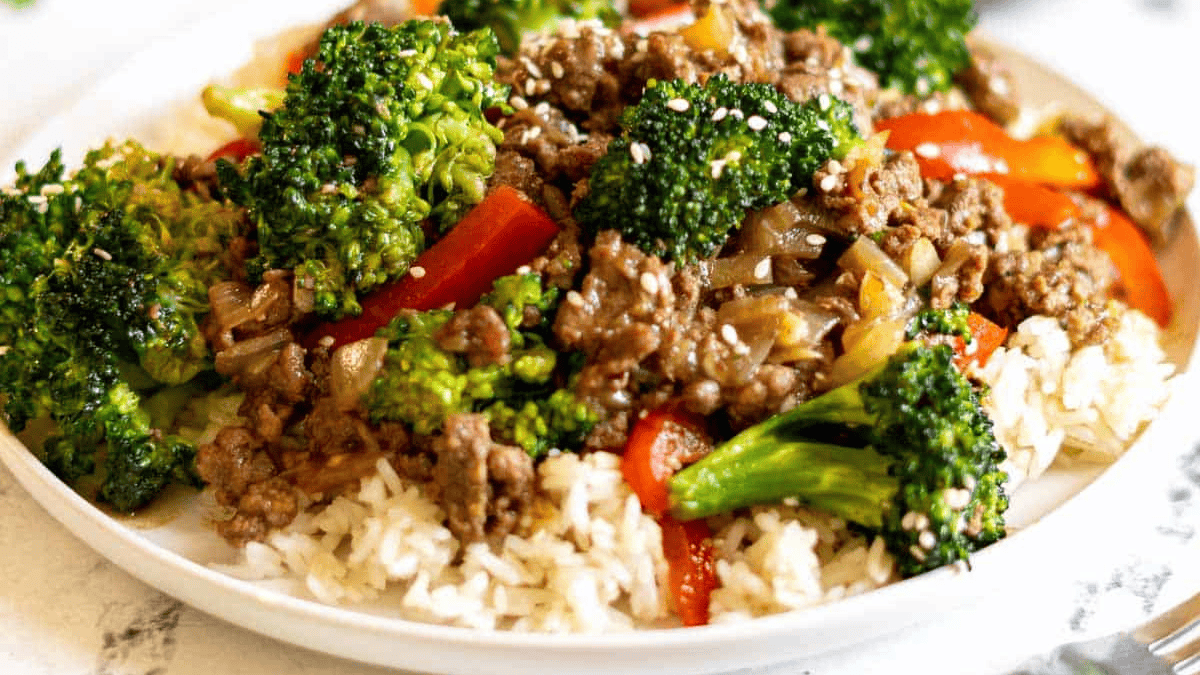 A plate of white rice topped with stir-fried ground beef, broccoli, red bell peppers, onions, and sprinkled with sesame seeds.