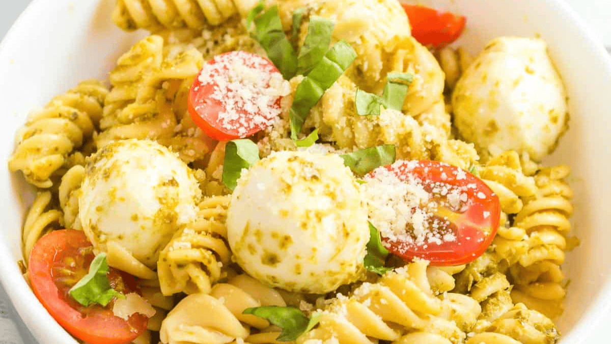 A bowl of pasta salad with rotini and shell pasta, mozzarella balls, halved cherry tomatoes, fresh basil, and pesto sauce, garnished with grated Parmesan cheese.