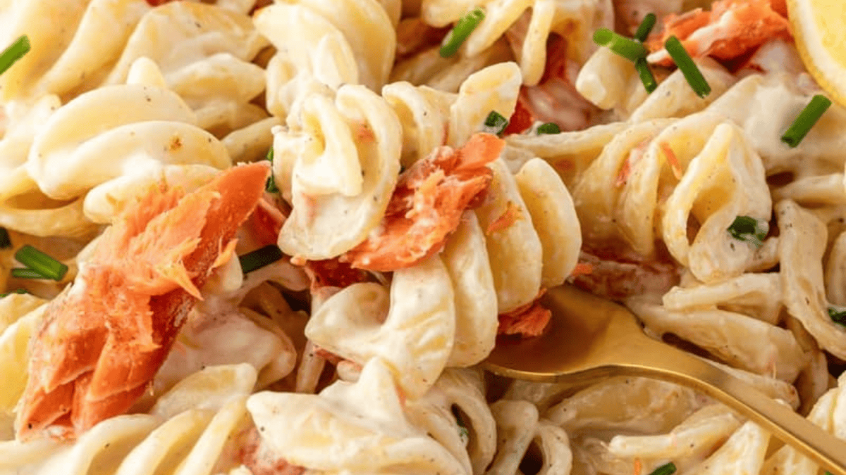 Close-up of creamy rotini pasta mixed with flakes of smoked salmon, garnished with chopped chives, and a gold fork twirling some pasta.