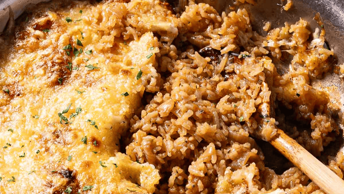 Close-up of a skillet with golden-brown baked chicken and seasoned rice, topped with melted cheese and garnished with chopped parsley. A wooden spoon is mixing the rice.