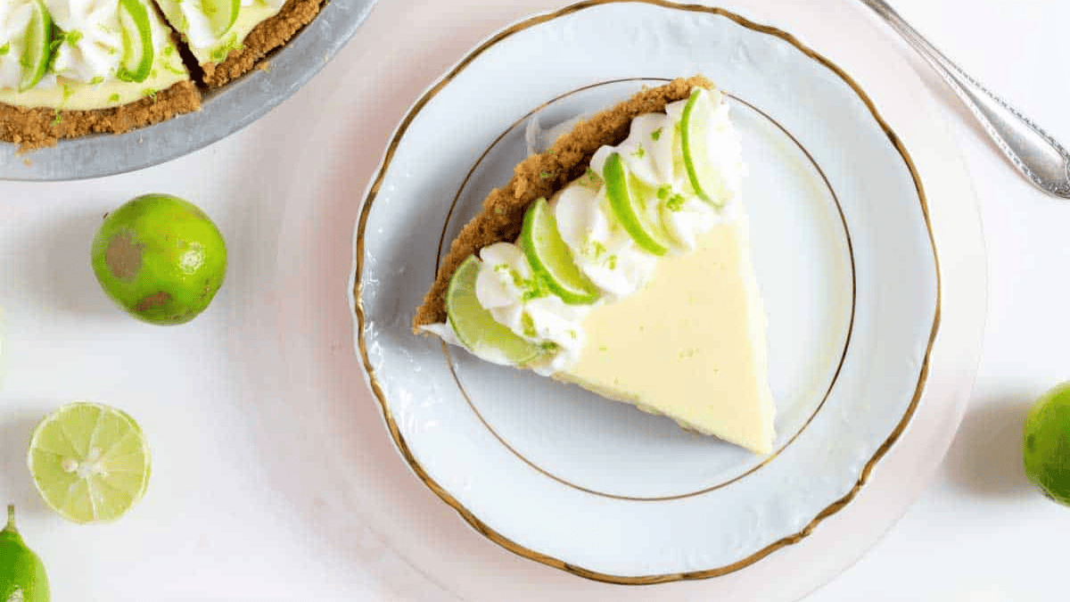 A slice of key lime pie with a graham cracker crust, topped with whipped cream and lime slices, on a white plate with a gold rim. Whole and halved limes are scattered nearby.