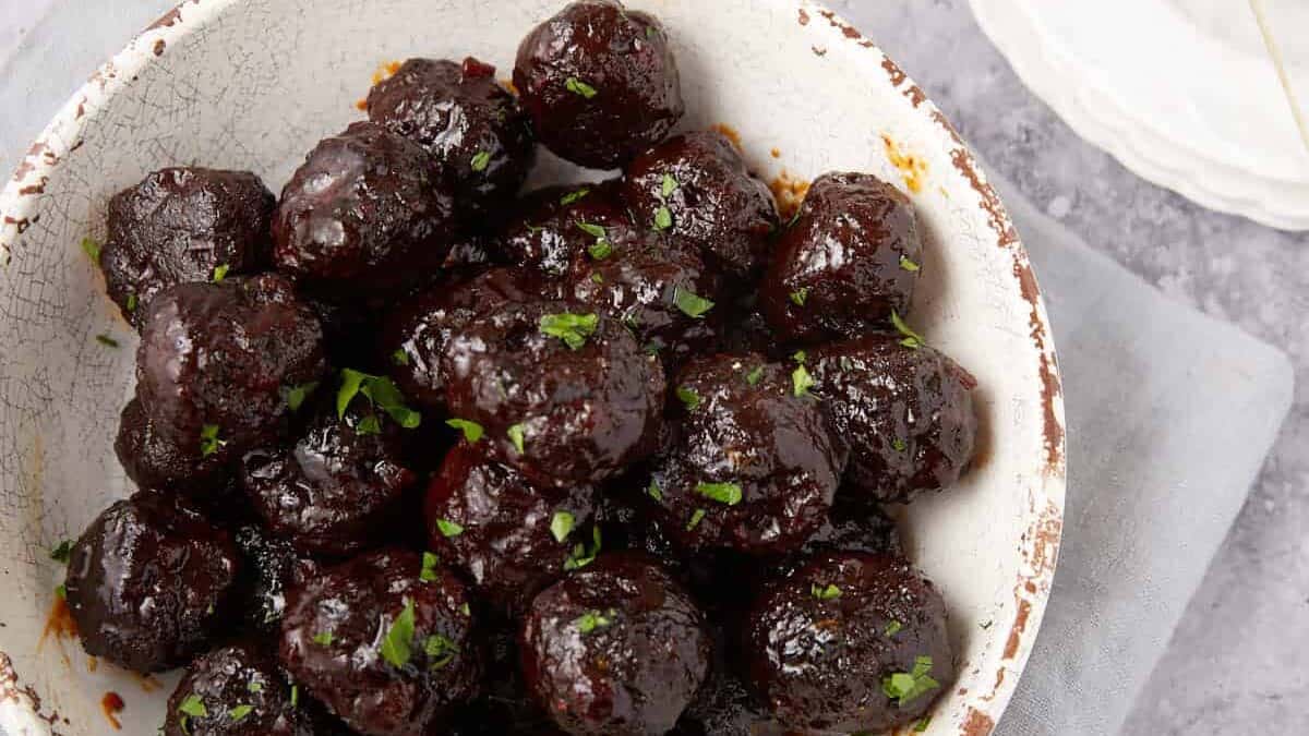 A white bowl filled with glazed Grape Jelly Meatballs garnished with chopped herbs sits on a light gray napkin. Nearby, a small white plate holds two more meatballs with cocktail sticks, and a small metal pot with sprouts is visible in the background.