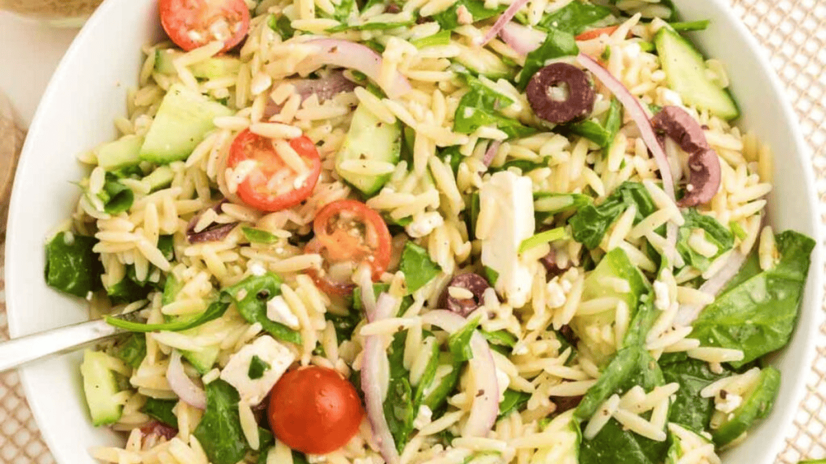 A bowl of orzo pasta salad with cherry tomatoes, cucumber, spinach, red onion, Kalamata olives, and feta cheese, mixed together and served with a fork.