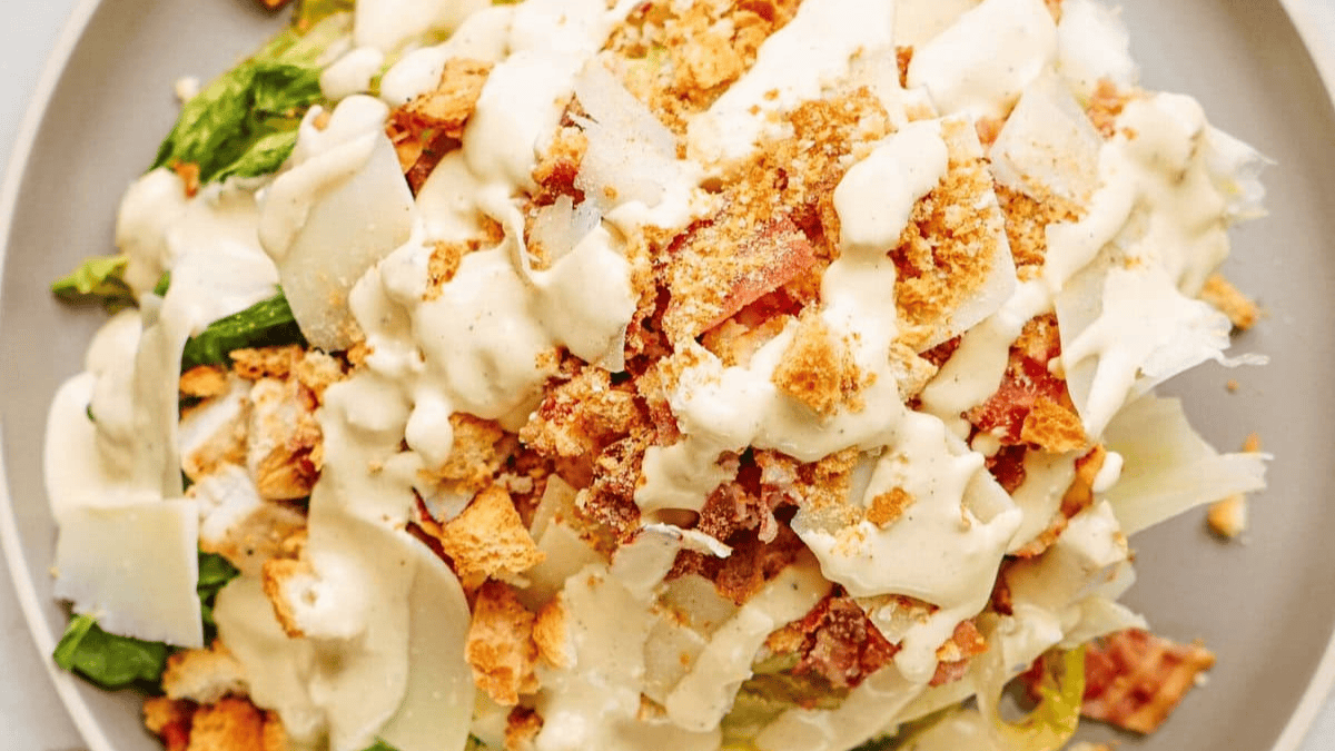 A close-up of a Caesar salad topped with creamy dressing, croutons, shredded Parmesan cheese, and chopped romaine lettuce on a light-colored plate.