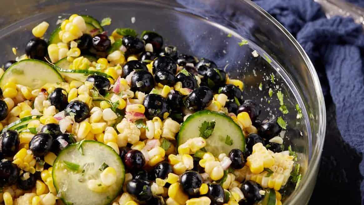 A clear glass bowl of corn salad with blueberries.