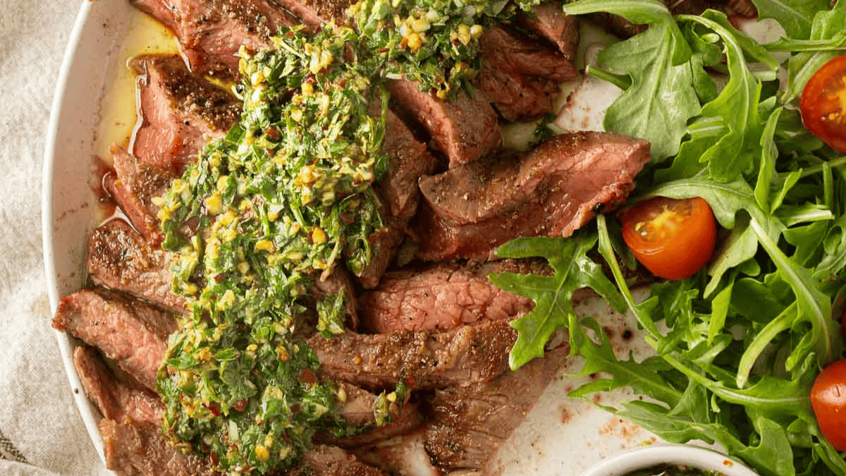 Sliced grilled steak topped with green chimichurri sauce, served alongside fresh arugula and cherry tomato salad on a white plate.