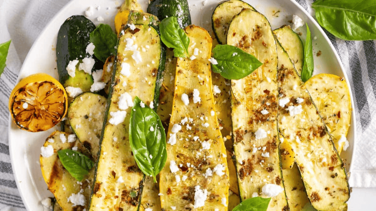 A plate of grilled zucchini and yellow squash slices topped with crumbled feta cheese, fresh basil leaves, and spices. A grilled lemon half sits on the side for garnish.