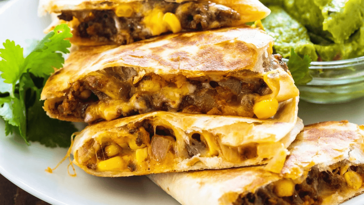 Four quesadilla slices filled with melted cheese, corn, onions, and seasoned meat are stacked on a plate. Fresh cilantro and a bowl of green guacamole are on the side.