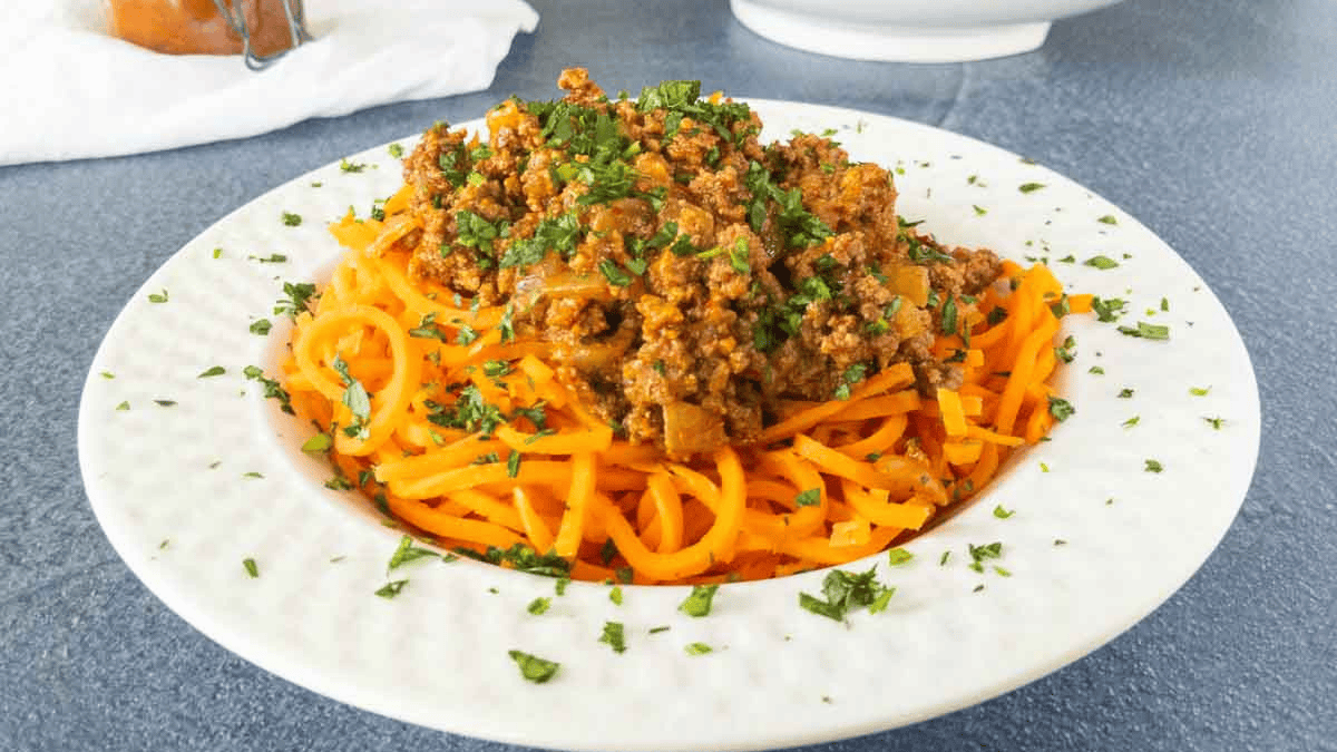 A white plate with spiralized carrot noodles topped with ground meat sauce and garnished with chopped parsley, set on a gray surface.