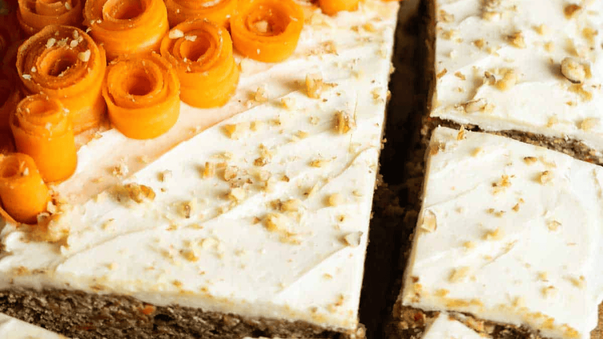 A close-up of a sliced carrot cake topped with cream cheese frosting, sprinkled with chopped nuts, and decorated with orange carrot ribbons on one corner.