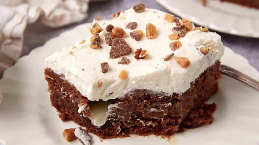 A slice of chocolate poke cake topped with whipped cream and toffee bits sits on a white plate, with a fork beside it and a creamy filling visible inside the cake.