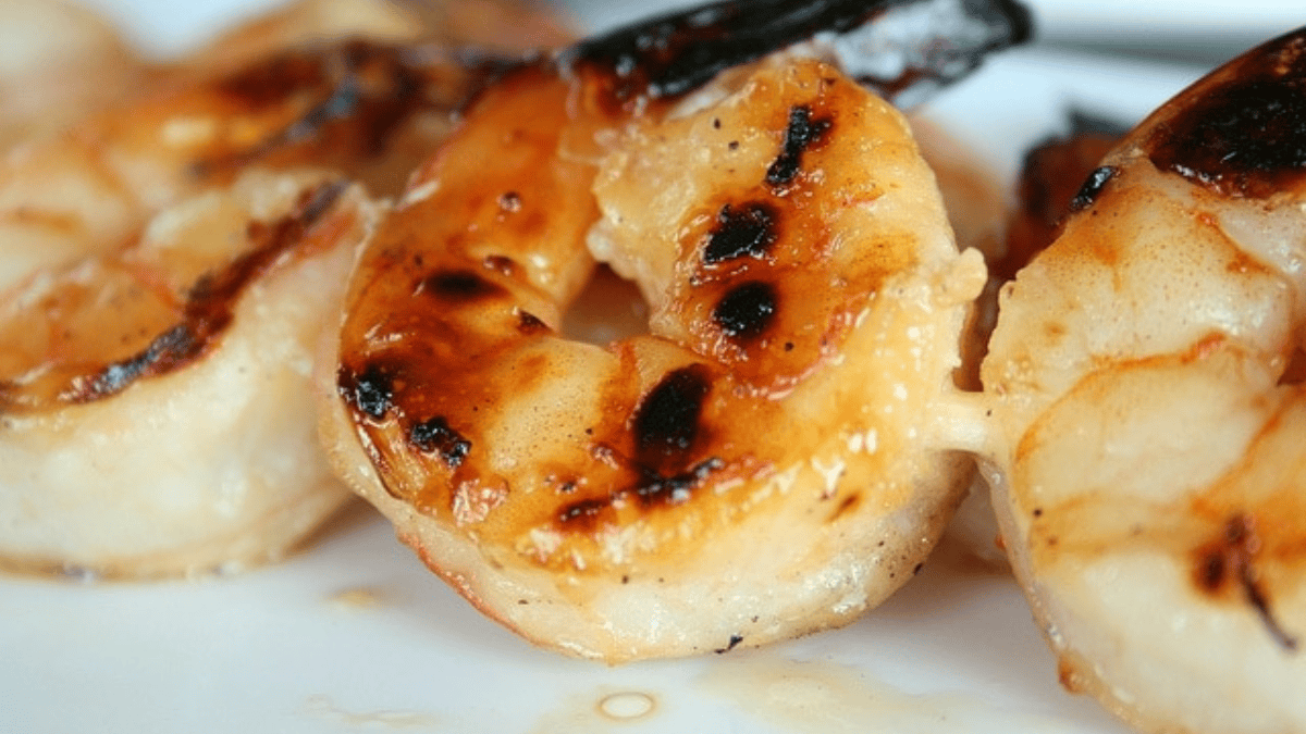 Close-up of grilled shrimp with a golden, slightly charred exterior, arranged on a white plate. The shrimp appear juicy and freshly cooked, with visible grill marks.
