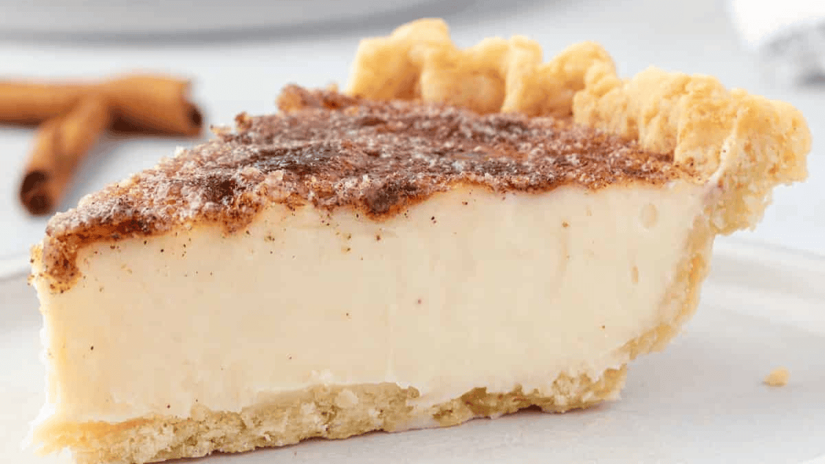 A close-up of a slice of creamy custard pie with a flaky golden crust and a cinnamon-sugar topping, placed on a white plate. Cinnamon sticks are blurred in the background.
