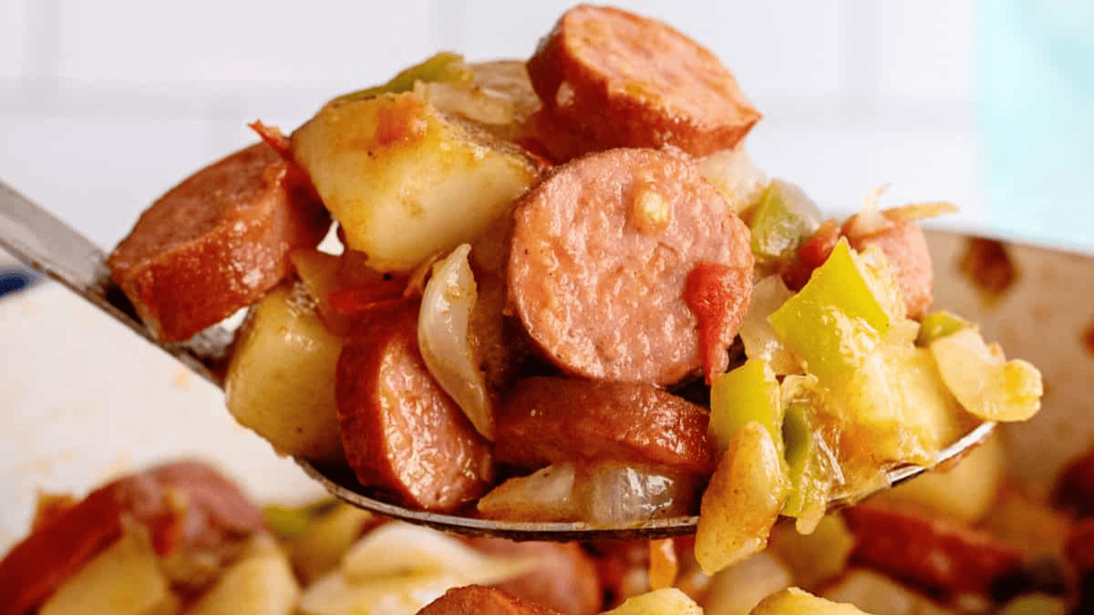 A close-up of a spoonful of sausage, potatoes, onions, and peppers, cooked together and seasoned, with more of the dish visible in the background.