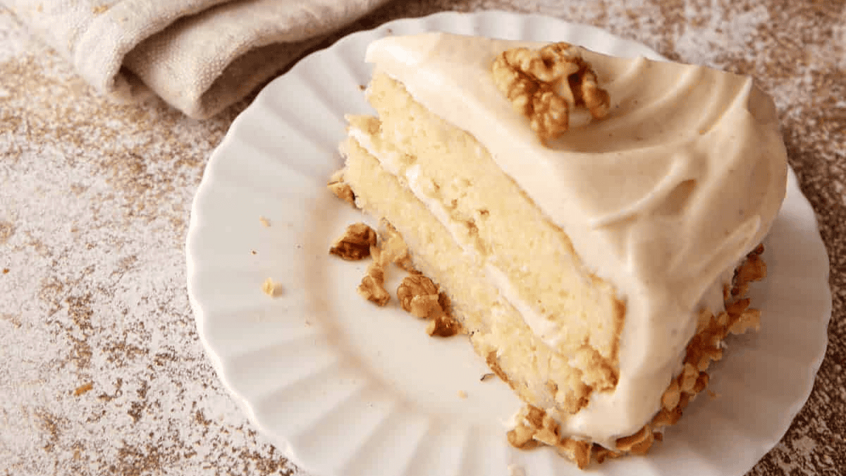 A slice of layered vanilla cake with creamy frosting and walnut topping on a white plate, with more walnuts around the base; a beige napkin is in the background.