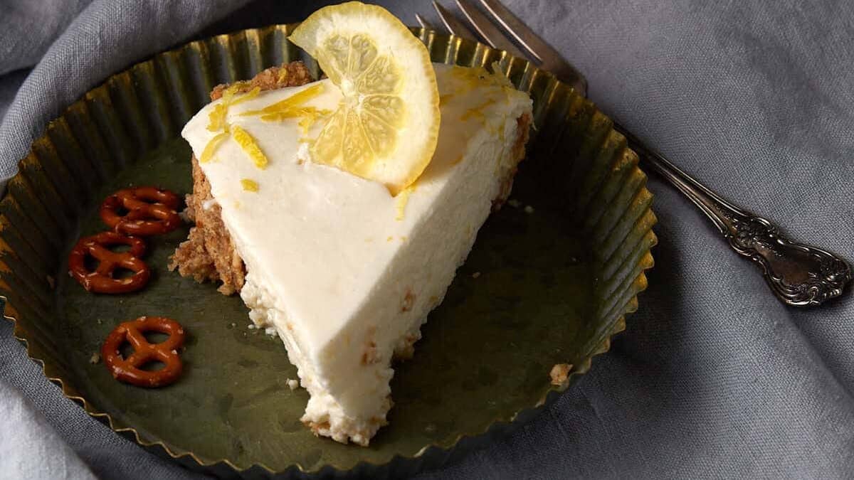 A slice of creamy lemon cheesecake garnished with a lemon slice and zest, served on a round metal plate with a few mini pretzels, next to a vintage fork on a gray fabric background.