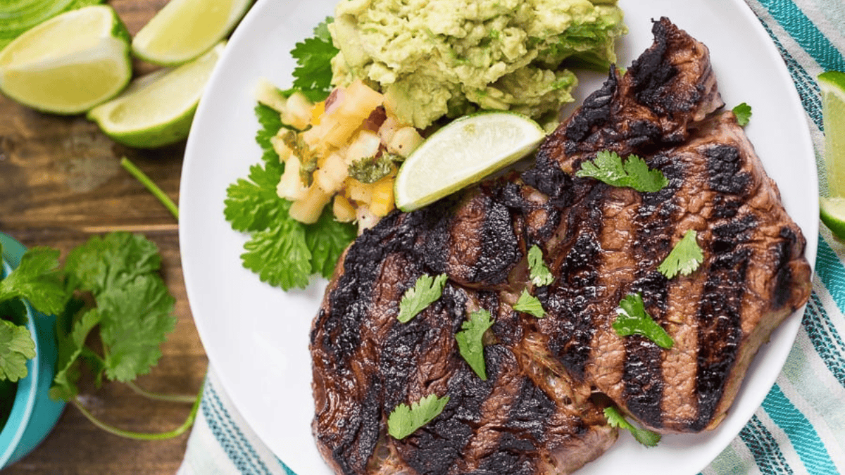 A plate with two grilled steaks garnished with cilantro, served with a scoop of guacamole, a lime wedge, and a small portion of salsa. Lime slices and cilantro are in the background.