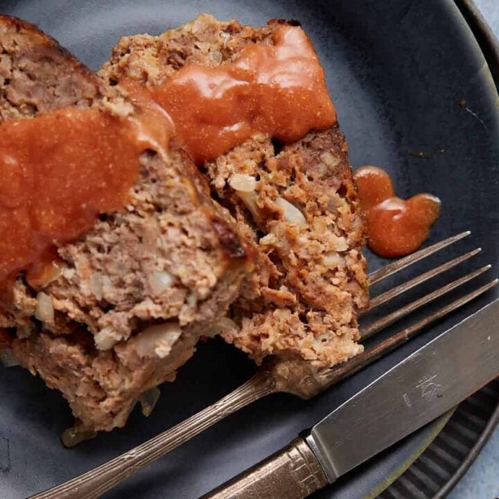 A close-up of two slices of savory meatloaf topped with a rich red sauce on a dark blue plate. A fork and knife are placed next to the meatloaf on the plate. The background surface is light blue, and a pumpkin-shaped dish is partially visible in the top right corner.