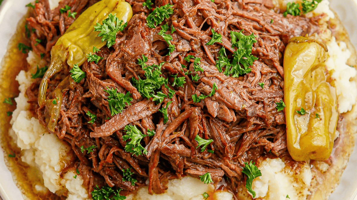 Shredded pot roast served over mashed potatoes, garnished with fresh parsley and whole pepperoncini peppers.