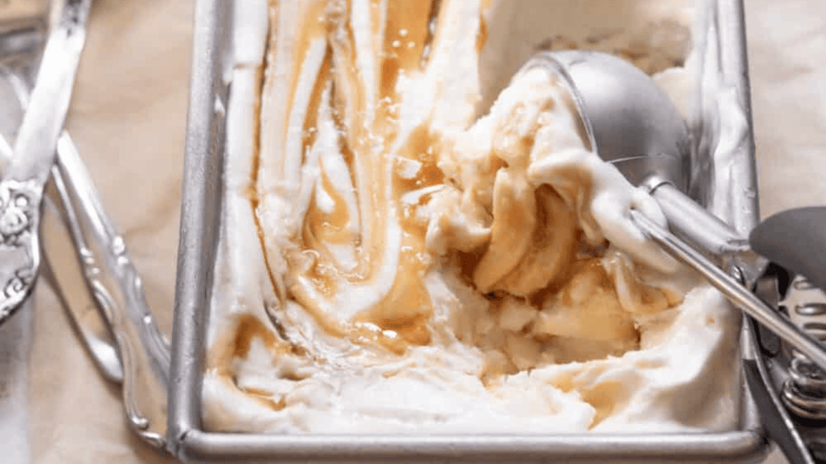 A metal ice cream scoop rests in a rectangular metal container filled with creamy vanilla ice cream swirled with caramel sauce. Some ice cream has been scooped out.