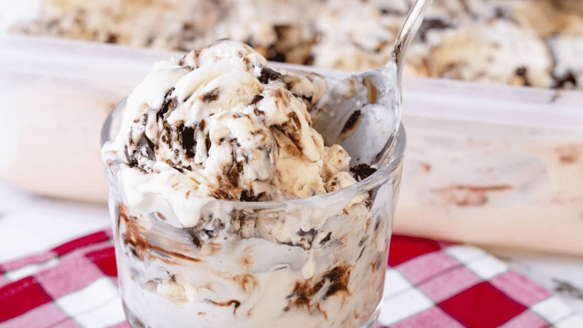 A glass bowl filled with creamy ice cream swirled with chocolate, set on a red and white checkered cloth, with a spoon in the bowl and an ice cream container in the background.