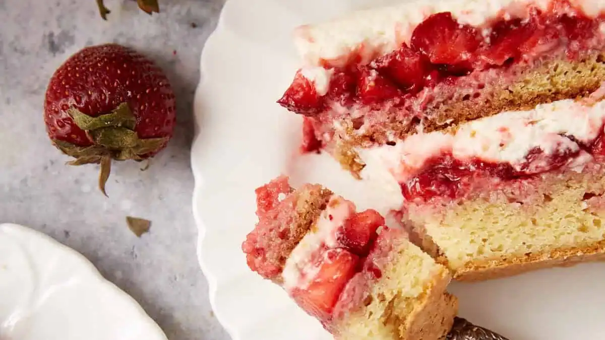 A close-up of a partially sliced strawberry shortcake on a white plate, showing layers of yellow cake, whipped cream, and strawberries. A whole strawberry sits beside the plate.