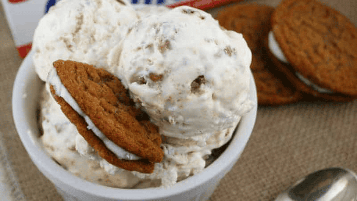 A white bowl filled with scoops of ice cream, featuring chunks of cookies and a sandwich cookie on top. More sandwich cookies and a spoon are visible in the background on a brown fabric surface.