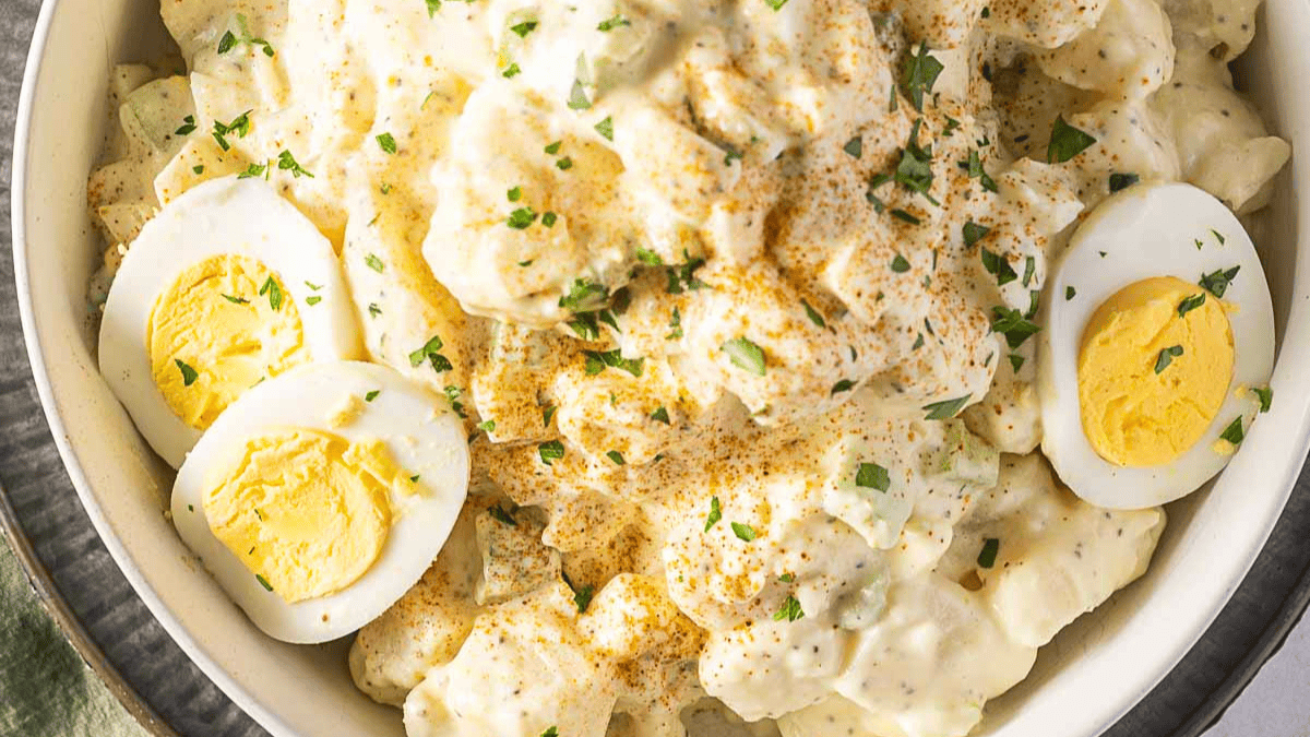 A bowl of creamy potato salad topped with chopped herbs and paprika, garnished with slices of hard-boiled egg.
