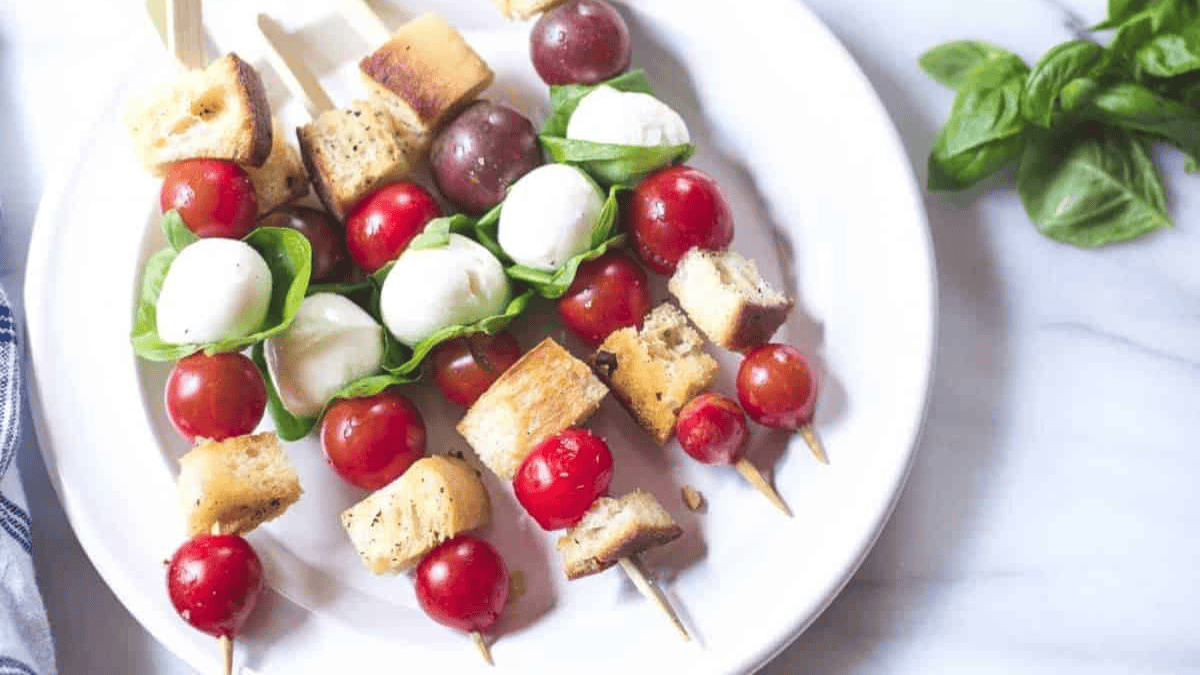 Three skewers with cherry tomatoes, mozzarella balls, fresh basil leaves, red grapes, and toasted bread cubes are arranged on a white plate, with a bunch of basil leaves nearby.