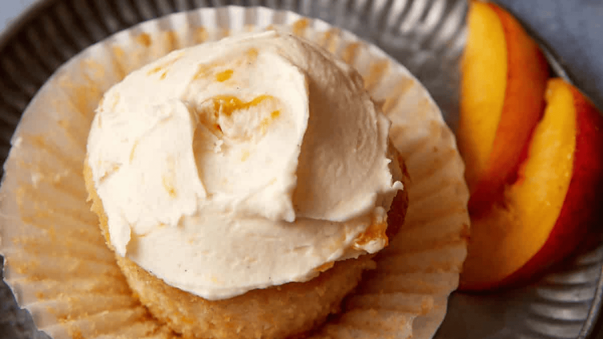 A cupcake with creamy frosting sits on a silver plate with its wrapper peeled back, next to two slices of fresh peach.