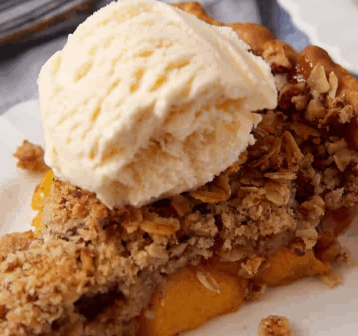 A slice of peach perfect crumble pie topped with a scoop of vanilla ice cream sits on a white plate. The crumbly oat topping and juicy filling make it one of our most irresistible recipes.