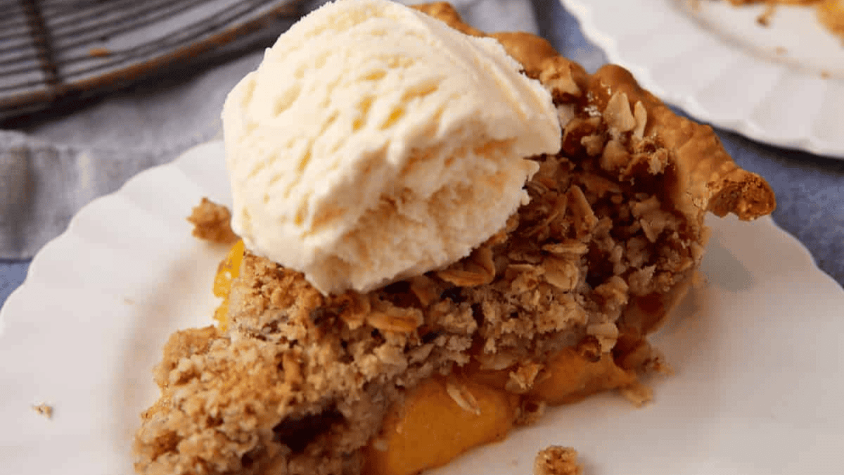 A slice of peach perfect crumble pie topped with a scoop of vanilla ice cream sits on a white plate. The crumbly oat topping and juicy filling make it one of our most irresistible recipes.