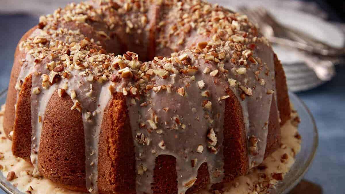 A bundt cake topped with a light glaze and chopped pecans, displayed on a glass cake stand with a stack of white plates and utensils in the background.