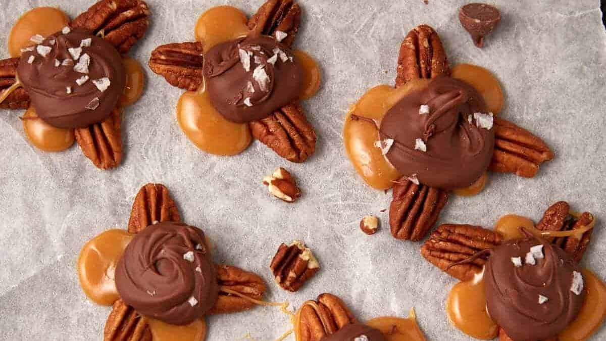 Clusters of pecans topped with caramel and swirled chocolate, some sprinkled with flaky sea salt, are arranged on parchment paper. One chocolate drop and pecan pieces are scattered around.
