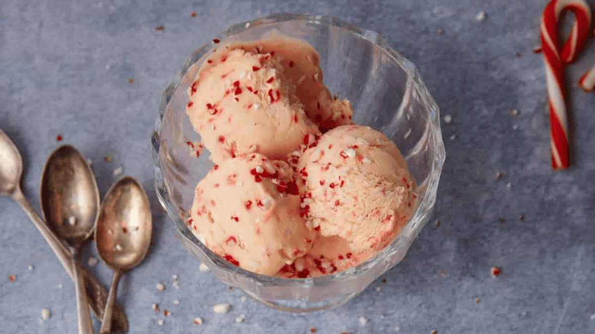 A glass bowl with three scoops of ice cream topped with crushed peppermint candy, placed on a blue surface next to three spoons and a candy cane.