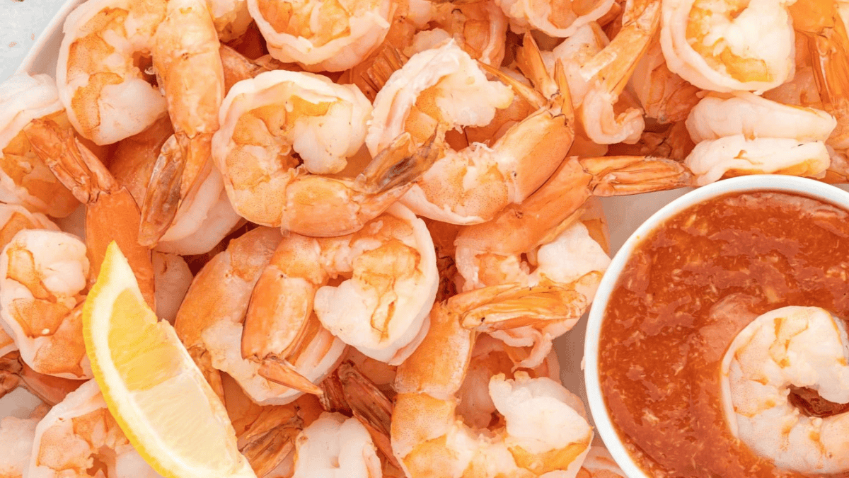 A plate of cooked shrimp with a wedge of lemon and a bowl of cocktail sauce for dipping.