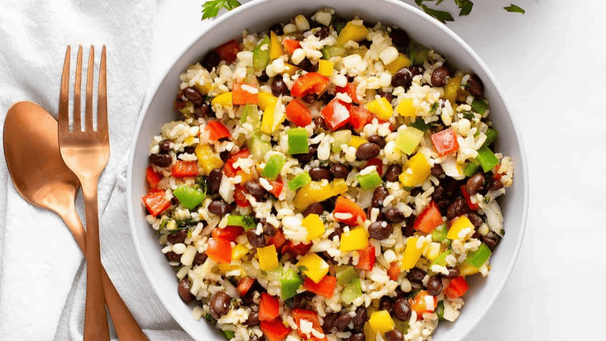 A bowl of southwestern-style rice salad with black beans, corn, and chopped red, yellow, and green bell peppers, placed on a white surface next to a fork and spoon.