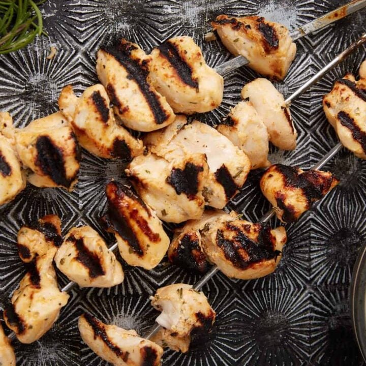 Grilled Ranch Chicken skewers with char marks on a textured dark surface, served beside a bowl of creamy dipping sauce and a small sprig of greenery in the corner.