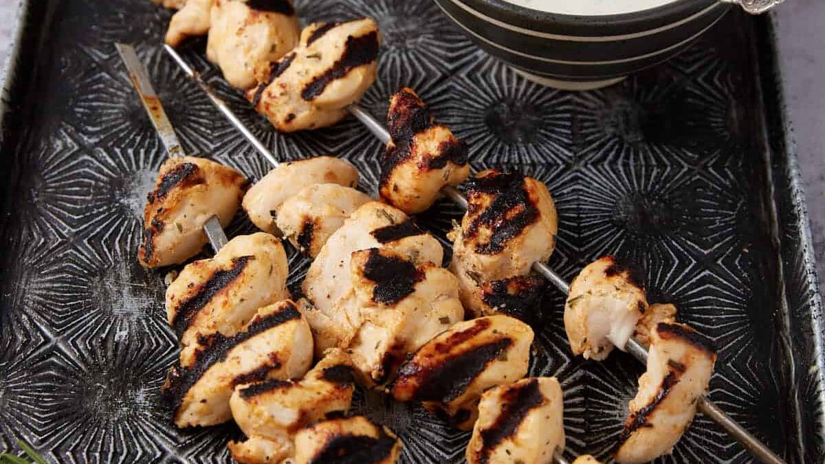 Grilled Ranch Chicken skewers on a patterned tray, served with a bowl of white dipping sauce and a stack of pita bread in the background. Fresh dill sprigs are placed beside the skewers.
