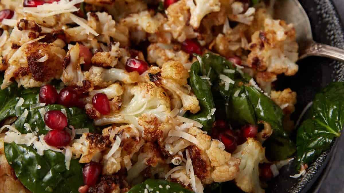 A bowl of roasted cauliflower mixed with fresh spinach leaves, topped with shredded cheese and pomegranate seeds, with a spoon resting in the bowl. A small bowl of grated cheese is visible in the background.