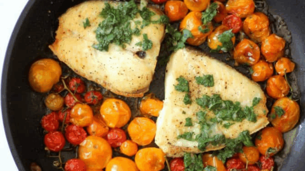 Two fillets of cooked fish topped with chopped herbs sit in a skillet surrounded by roasted red and yellow cherry tomatoes.
