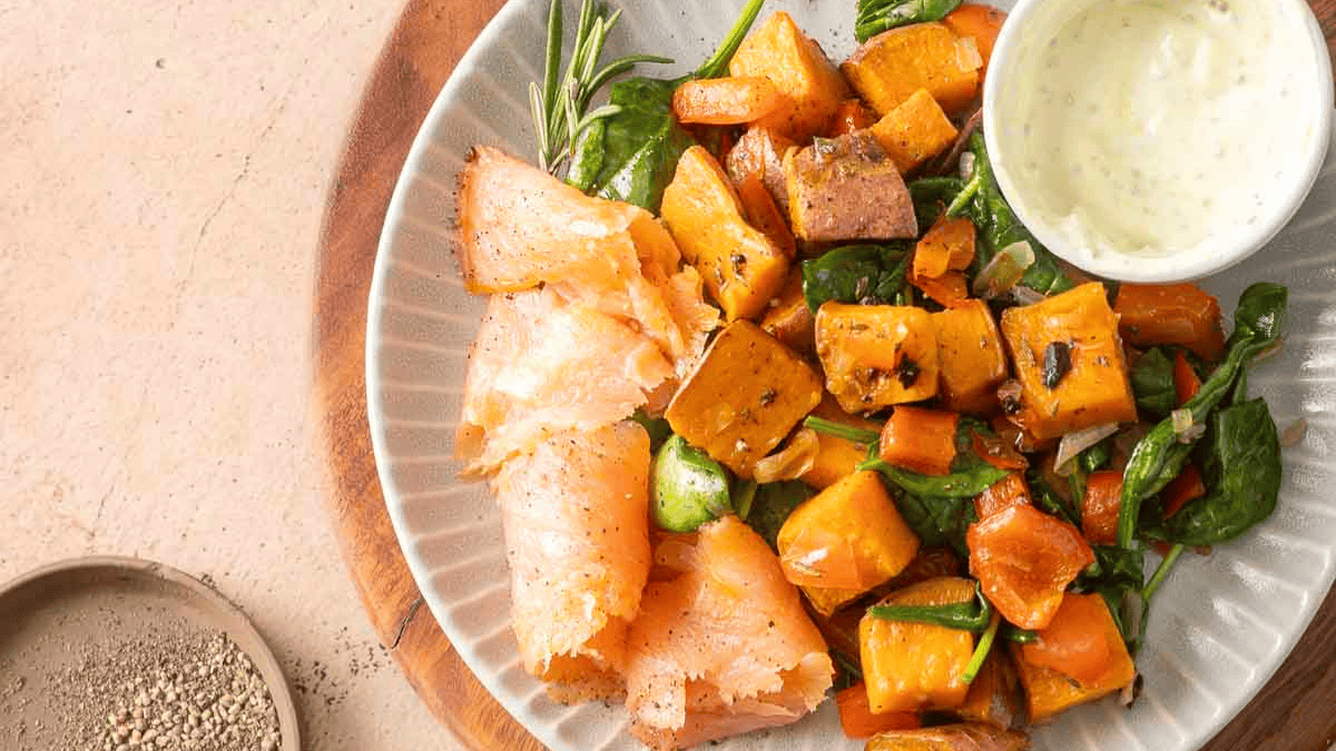 A plate with slices of smoked salmon, roasted sweet potato and vegetable salad, a sprig of rosemary, and a small bowl of creamy sauce. A dish of ground pepper sits nearby on the table.