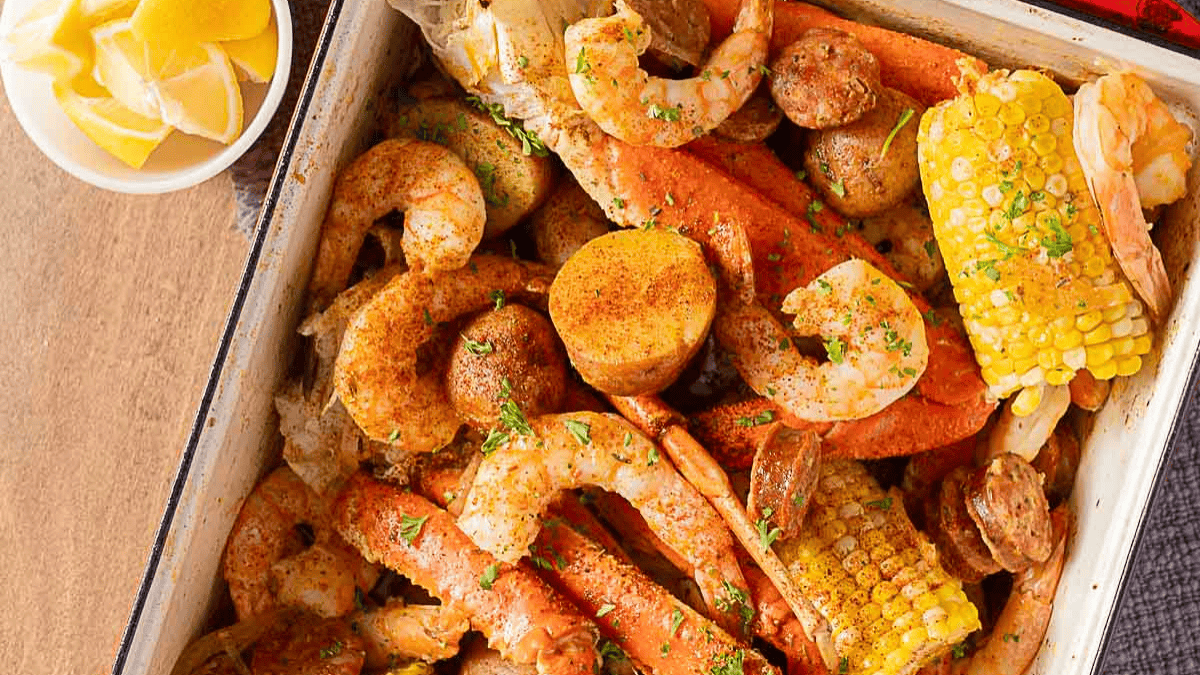 A tray filled with seasoned shrimp, crab legs, corn on the cob, potatoes, and sausage slices, garnished with parsley. A bowl of lemon wedges sits nearby on the table.